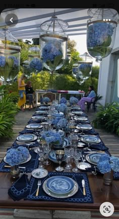 a long table is set with blue flowers and place settings for an outdoor dinner party