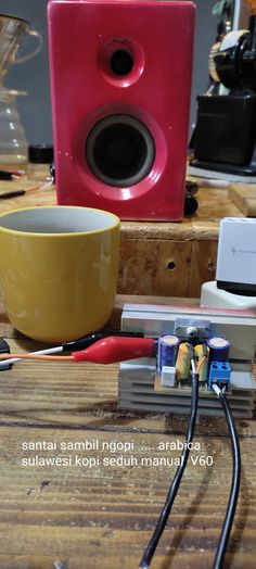 a red speaker sitting on top of a wooden table next to a yellow cup and wires