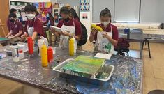 three girls in red shirts and white masks are painting with yellow tubes on a table