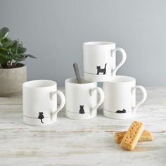 four coffee mugs with black cats on them next to crackers and a potted plant