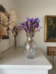 two vases with purple flowers sitting on a white table in front of a painting