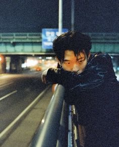 a man leaning on a rail in front of a highway at night with traffic lights behind him