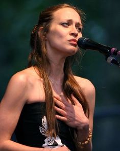 a woman with long hair standing in front of a microphone and holding her hands to her chest