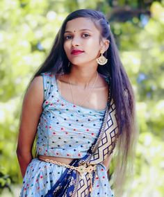 a woman with long hair wearing a blue sari and gold jewelry, standing in front of trees