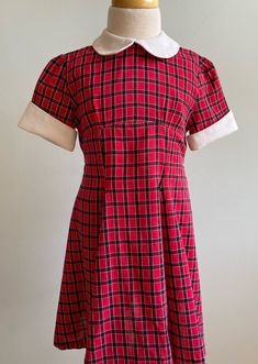 "This dress has a very prim and proper look to it and is just incredibly charming when worn. Made at home without a label, the fabric is a cotton dobby with a red ground and a black and white check pattern woven into it. The front bodice is high and shaped by the tiniest of pleats. The skirt is straight through midriff, then gored (A-line panels seamed together). This gives a nipped in waist and very twirly skirt look to the dress. Sleeves are short with just the hint of a gather at crown. Cuffs Red Short Sleeve School Dress, Retro Cotton Plaid Dress, Fitted Red Plaid Dress With Short Sleeves, Red Fitted Plaid Dress With Short Sleeves, Retro Plaid Cotton Dress, Retro Fitted Dress For School, Retro Cotton School Dress, Retro Fitted School Dress, Classic Red Cotton Dress