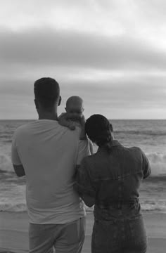 two adults and a baby are standing on the beach looking at the waves in the ocean