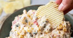 a hand holding a potato chip over a bowl of corn salad with ranch dressing in the background