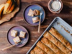 two plates with pastries on them next to a pan filled with bananas and cinnamon rolls