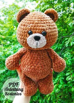 a crocheted brown teddy bear sitting on top of someone's hand with trees in the background