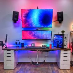 a computer desk with two monitors and speakers on top of it in front of a colorful wall