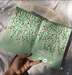 an open book with flowers and leaves on the pages is being held by someone's hand