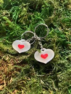 two heart shaped glass key chains sitting on top of green moss covered ground with red hearts hanging from them