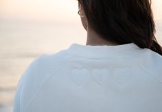 the back of a woman's white sweatshirt with three hearts on it, facing an ocean