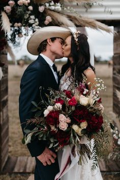 a bride and groom kissing in front of an arch decorated with flowers, feathers and foliage