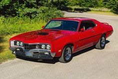 a red muscle car parked on the side of a road next to grass and trees