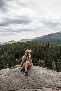 a woman sitting on top of a large rock talking on a cell phone while wearing a hat