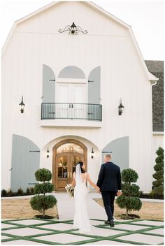 a bride and groom walking towards the front of a large white barn with blue shutters