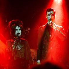 two people on stage with red lights behind them and one person in costume holding a microphone