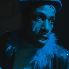 a man with blue paint on his face wearing a hat and feathered coat in the dark