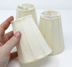 a hand is holding two small lampshades on a white tableclothed surface