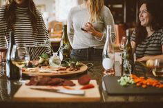 three women standing around a table with food and wine