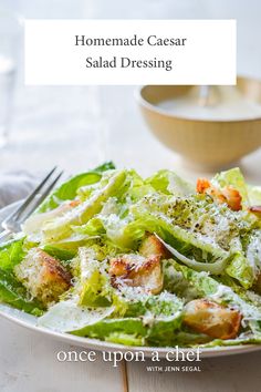 a white plate topped with salad next to a fork and bowl filled with dressing on top of a wooden table