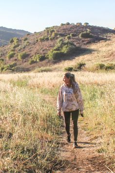 a woman walking down a dirt road next to a lush green hillside