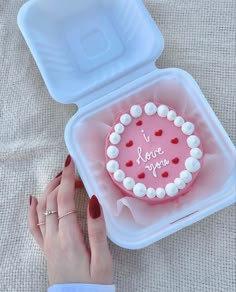 a woman's hand is holding a pink heart shaped cake in a plastic container