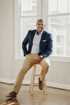 a man sitting on top of a wooden stool in front of a window wearing a blue jacket and khaki pants