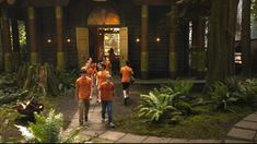 several people in orange shirts are walking towards a house surrounded by trees and ferns with lights on