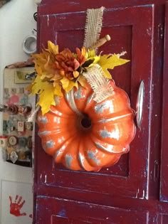 an orange pumpkin hanging from a red door with fall leaves on it's front