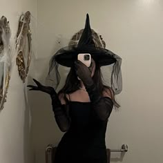 a woman in a black dress taking a selfie with her cell phone wearing a witch hat