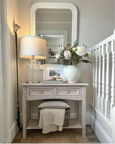 a white table with flowers and a mirror on it next to a stair case in a home