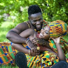 a man and woman are sitting on the ground in front of some trees, smiling at each other