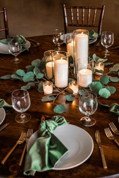 A wooden table set with gray china, green velvet napkins and gold flatware. In the center, a cluster of candles surrounded by eucalyptus fronds. Beautiful Dinner Party Table Settings, Round Holiday Table Setting, Meal Table Decoration, Dinner Table Setting Ideas Simple, Small Table Setting Ideas, Table Settings Dinner Party, Dinner Party Table Settings Casual, Corporate Holiday Party Decorations, Corporate Christmas Party Ideas