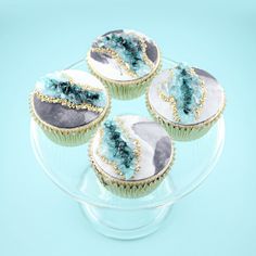 four cupcakes with blue frosting and gold decorations on a clear cake plate