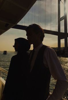 two people standing next to each other on a boat near the water with a bridge in the background