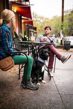 two people sitting at an outdoor table with a dog on the sidewalk next to them