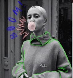 a woman in grey and green sweater blowing pink bubble into her mouth while standing next to a building