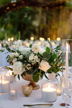 white flowers and greenery in a vase on a table with silverware, candles and plates