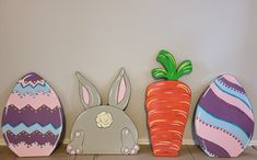 three painted easter eggs and two decorated carrots on the floor in front of a wall