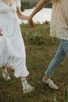 a man and woman holding hands while walking in the grass near a body of water