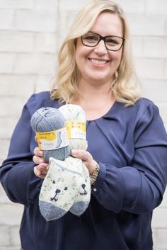 a woman holding two pairs of socks in front of a white brick wall smiling at the camera