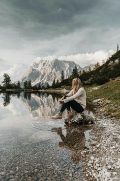 a woman sitting on the edge of a lake with her feet in the water and mountains in the background