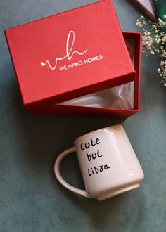 a white coffee mug with writing on it next to a red box and some baby's breath flowers