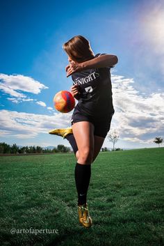 a young woman kicking a soccer ball on top of a green field with blue sky in the background