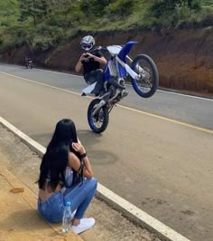 a woman sitting on the side of a road next to a man riding a motorcycle