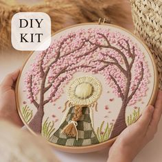 a person is holding a small embroidery kit with pink flowers on it and a tree in the background