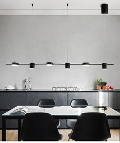 a dining table with black chairs and lights hanging from the ceiling above it, in front of a kitchen island