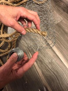 two hands are holding some rope on top of a wooden floor and another hand is tying it to a piece of metal wire